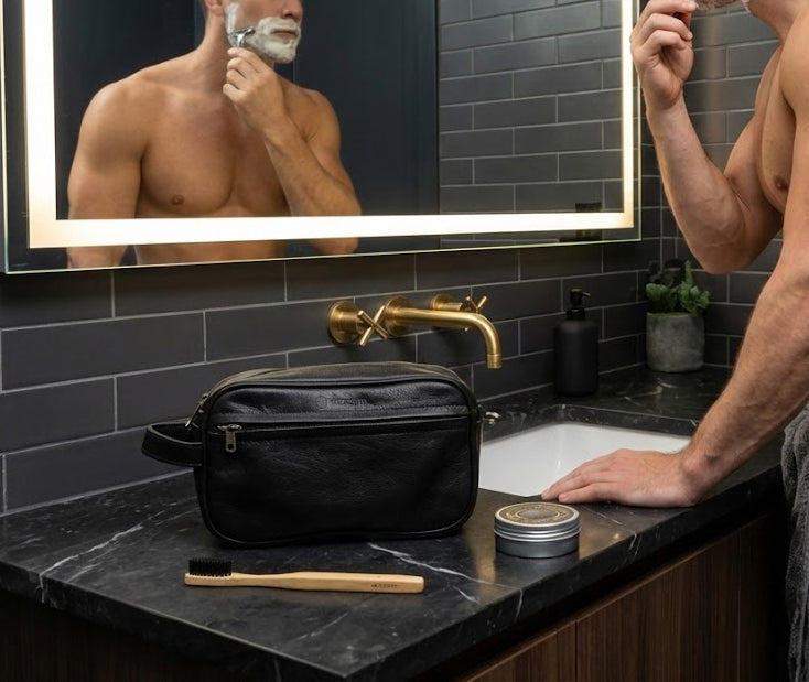Full-grain black leather RIDGE cosmetic bag on a bathroom counter, shown beside grooming items while a man shaves in the mirror. Premium handcrafted toiletry bag designed for travel and daily grooming essentials.
